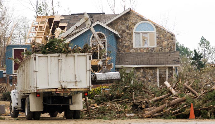 truck cleaning up fallen tree debris in front of a storm-damaged house